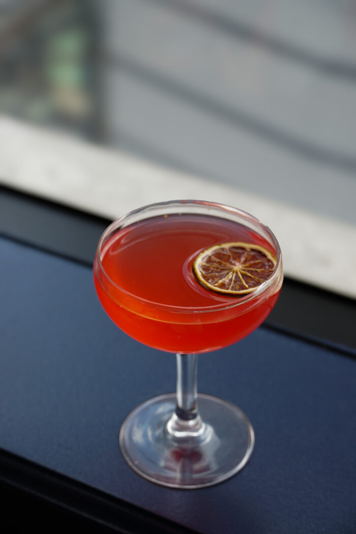Bright red lychee cosmopolitan served in a coupe glass with a dried citrus garnish.