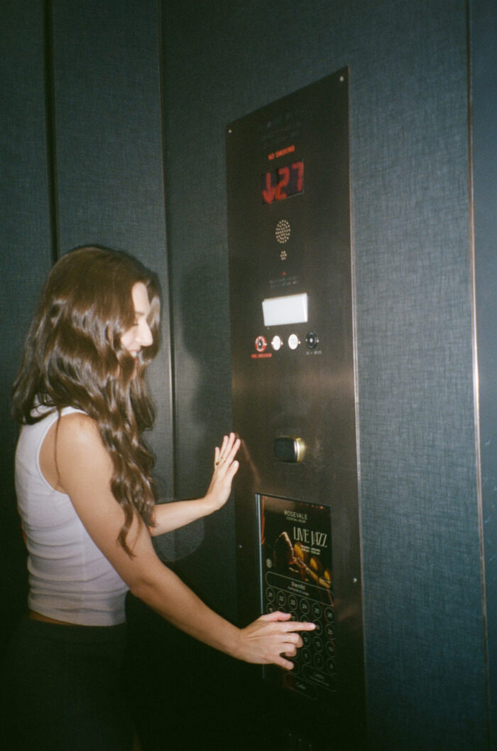 Woman in an elevator pressing a button panel, with ‘Live Jazz’ poster visible below the controls.