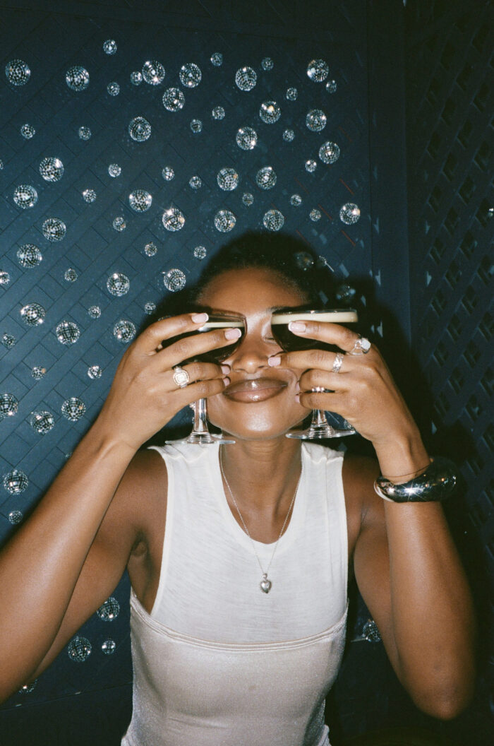 Smiling woman holding up two espresso martinis, playfully covering her eyes, against a wall of mirrored orbs.