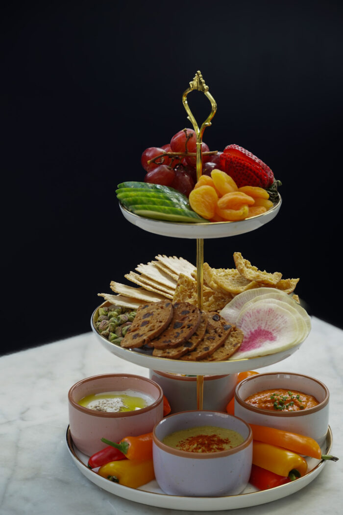 Three-tiered food tower with fruits, crackers, nuts, and colorful dips, displayed on a marble table.