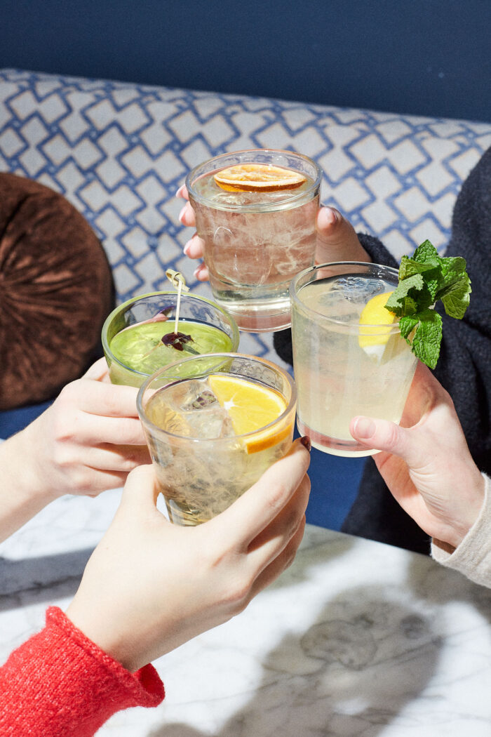 Four hands toasting with assorted cocktails over a marble table in a cozy lounge setting.