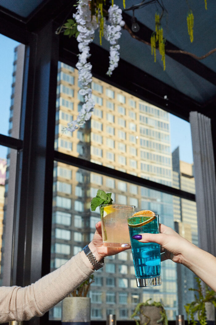Two people clinking colorful cocktails in a sunlit rooftop bar with high-rise buildings in the background.