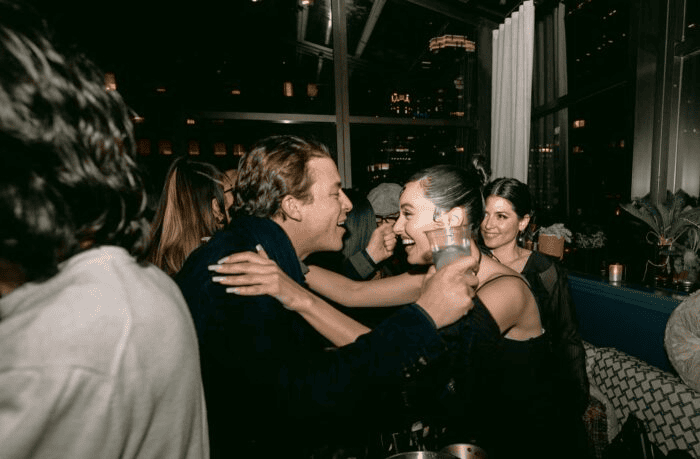 Man and woman smiling and embracing at a nighttime indoor party, surrounded by other guests and city lights in the background.