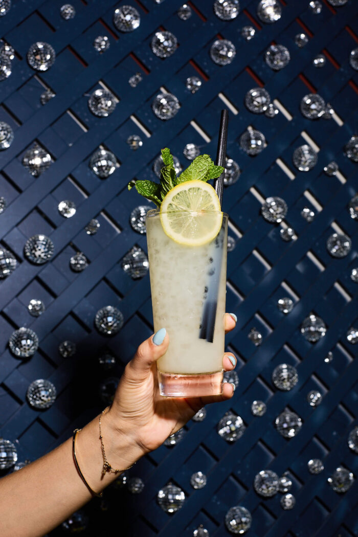 Hand holding a lemon and mint cocktail against a blue disco wall.