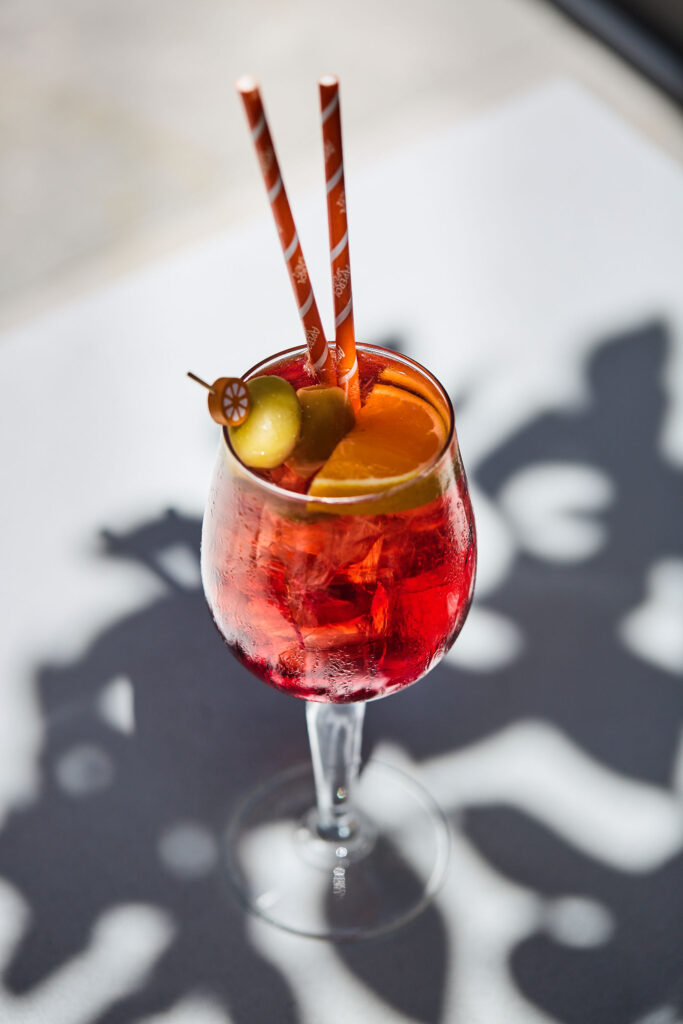 Bright red-orange cocktail with olive and citrus garnish, served in a stemmed glass with two striped straws.