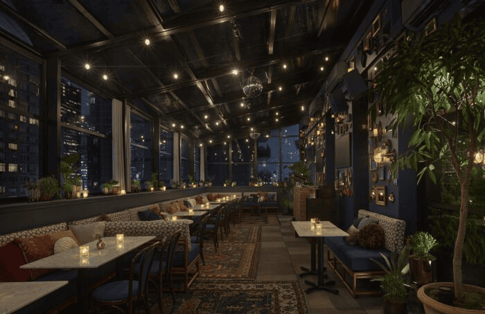 Elegant rooftop lounge at night with string lights, candlelit tables, lush plants, and city skyline views through large windows.
