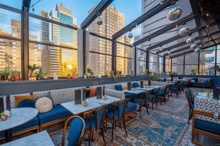 Chic rooftop lounge with floor-to-ceiling windows, patterned rugs, velvet seating, and city skyline views at sunset.