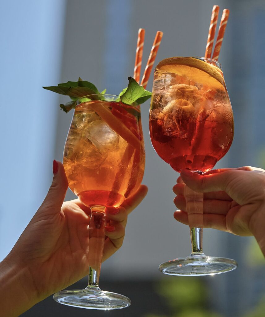 Two colorful spritz cocktails with citrus and straws held up for a toast in daylight.