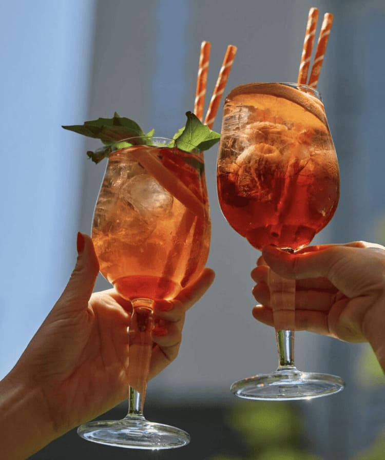 Two hands clink Aperol spritz cocktails garnished with orange slices and mint in front of a blurred cityscape.