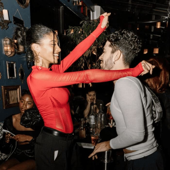 Two stylishly dressed people smiling and dancing closely in a dimly lit lounge, surrounded by others seated at a table with drinks.
