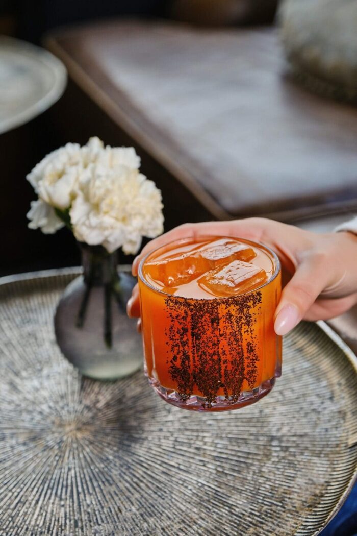 Woman holding a vibrant orange cocktail with chili rim on a silver tray beside white carnations.