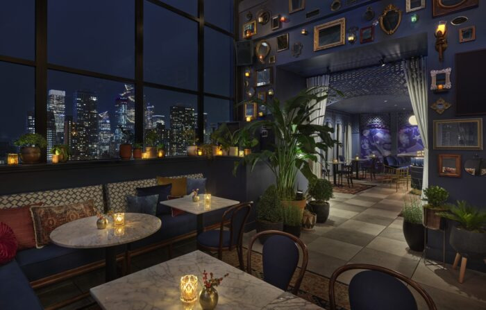 Best Rooftop Bar in Midtown NYC: Experience Starchild’s Skyline Views Elegant rooftop lounge with marble tables, cozy candle lighting, and panoramic nighttime views of the city skyline through tall windows. The interior features lush potted plants, picture frames on the wall, and a moody, upscale ambiance.