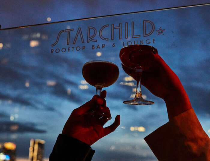 Best Rooftop Bar in Midtown NYC: Experience Starchild’s Skyline Views Two people toast with cocktails on a rooftop at night, with a scenic city skyline in the background and “Starchild Rooftop Bar & Lounge” written on the glass panel behind them.