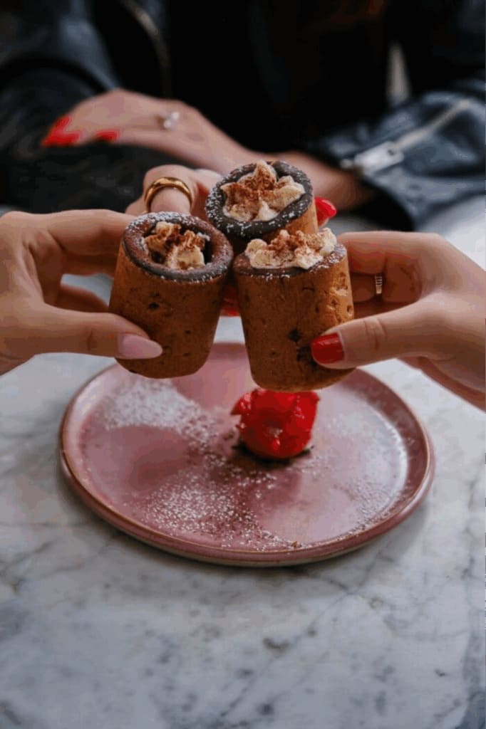 Three hands holding up dessert cookie shot glasses filled with whipped cream and dusted with cocoa, over a marble table with a red scoop of sorbet on a pink plate in the background.