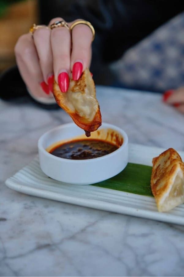 Hand with bright red nails dipping a crispy dumpling into a small bowl of dark sauce on a marble table.