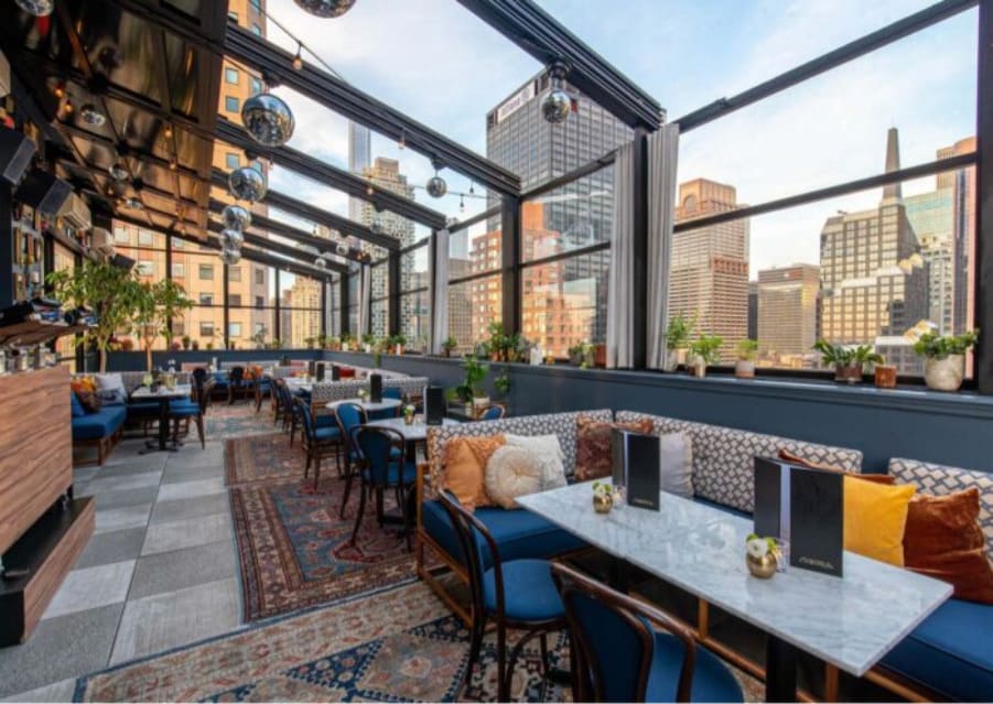 Best Rooftop Bar in Midtown NYC: Experience Starchild’s Skyline Views Enclosed rooftop lounge with marble tables, blue seating, and city skyline views through large glass windows.