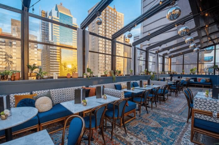 Chic rooftop lounge with floor-to-ceiling windows, patterned rugs, velvet seating, and city skyline views at sunset.