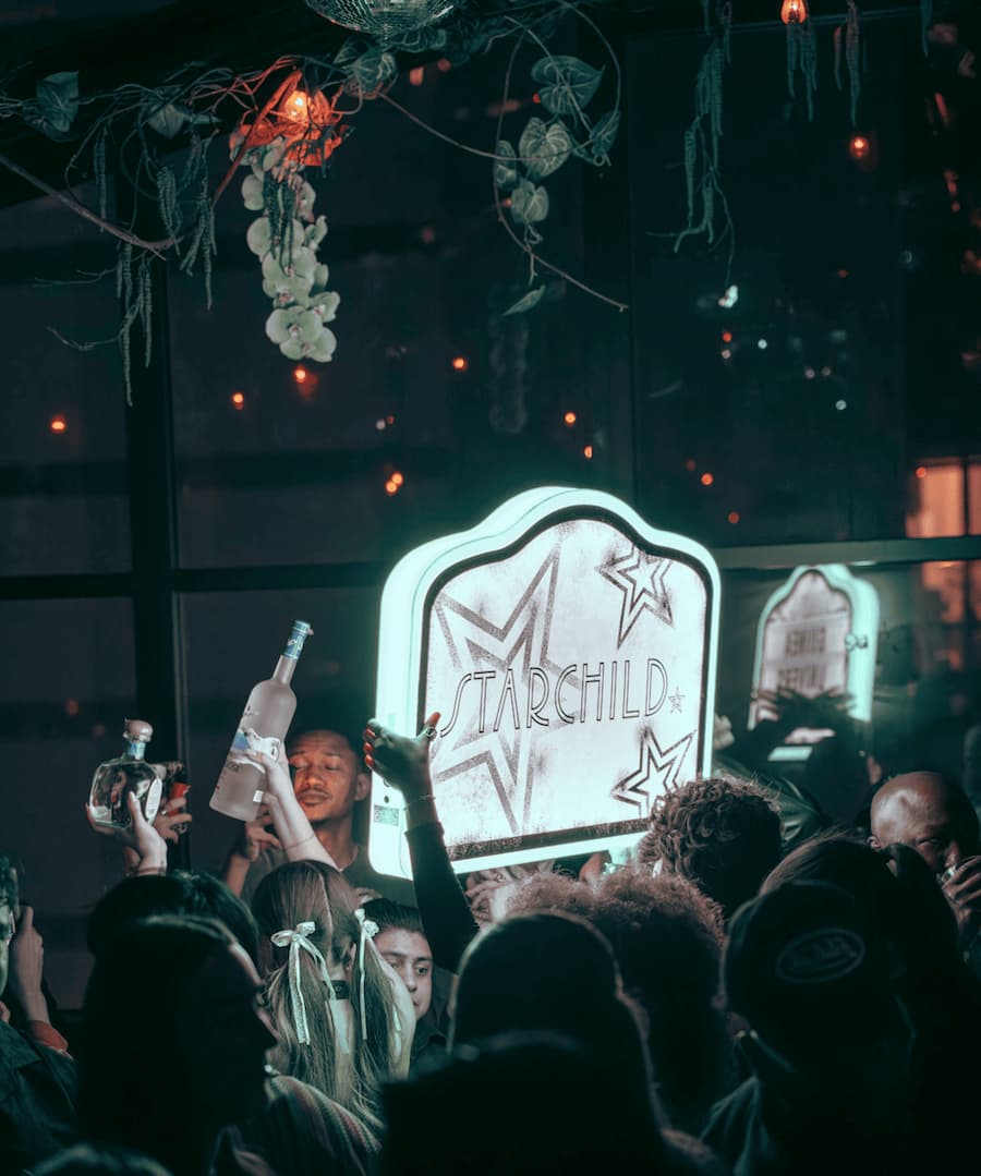 Crowded nightclub scene featuring guests celebrating around an illuminated Starchild sign while staff hold liquor bottles, with hanging floral decorations and dim ambient lighting.