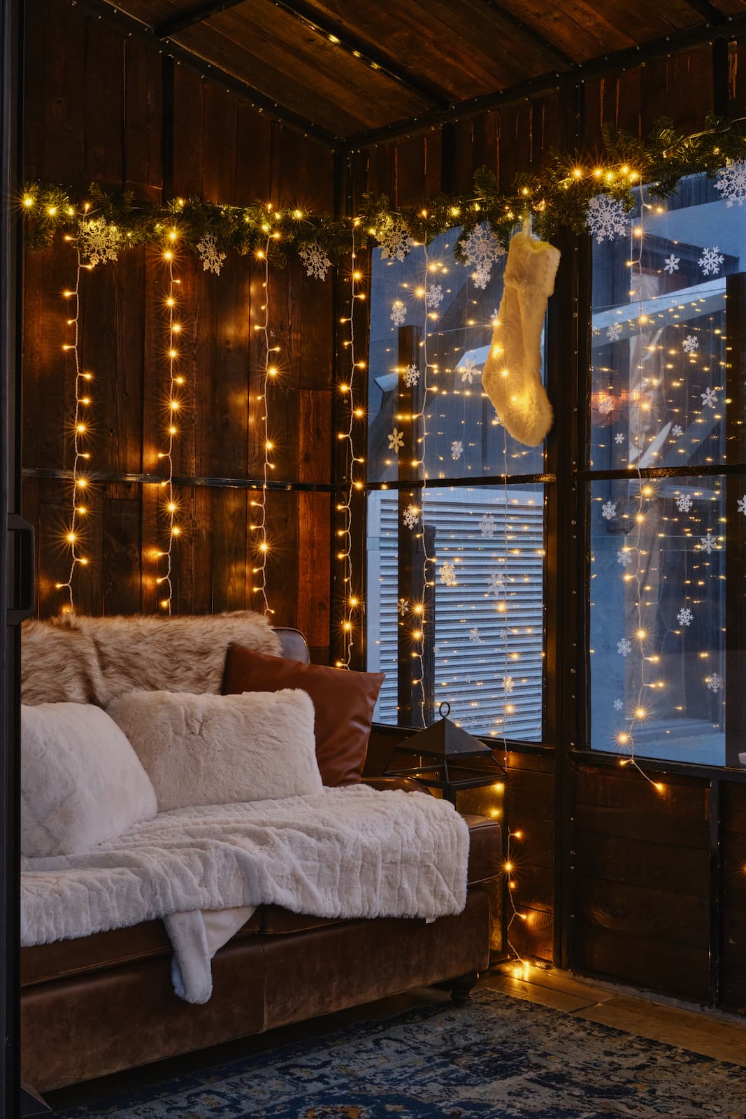 Cozy lounge with a sofa, plush blankets, warm string lights, hanging snowflakes, and a Christmas stocking inside a wood-paneled space at StarChild Rooftop.