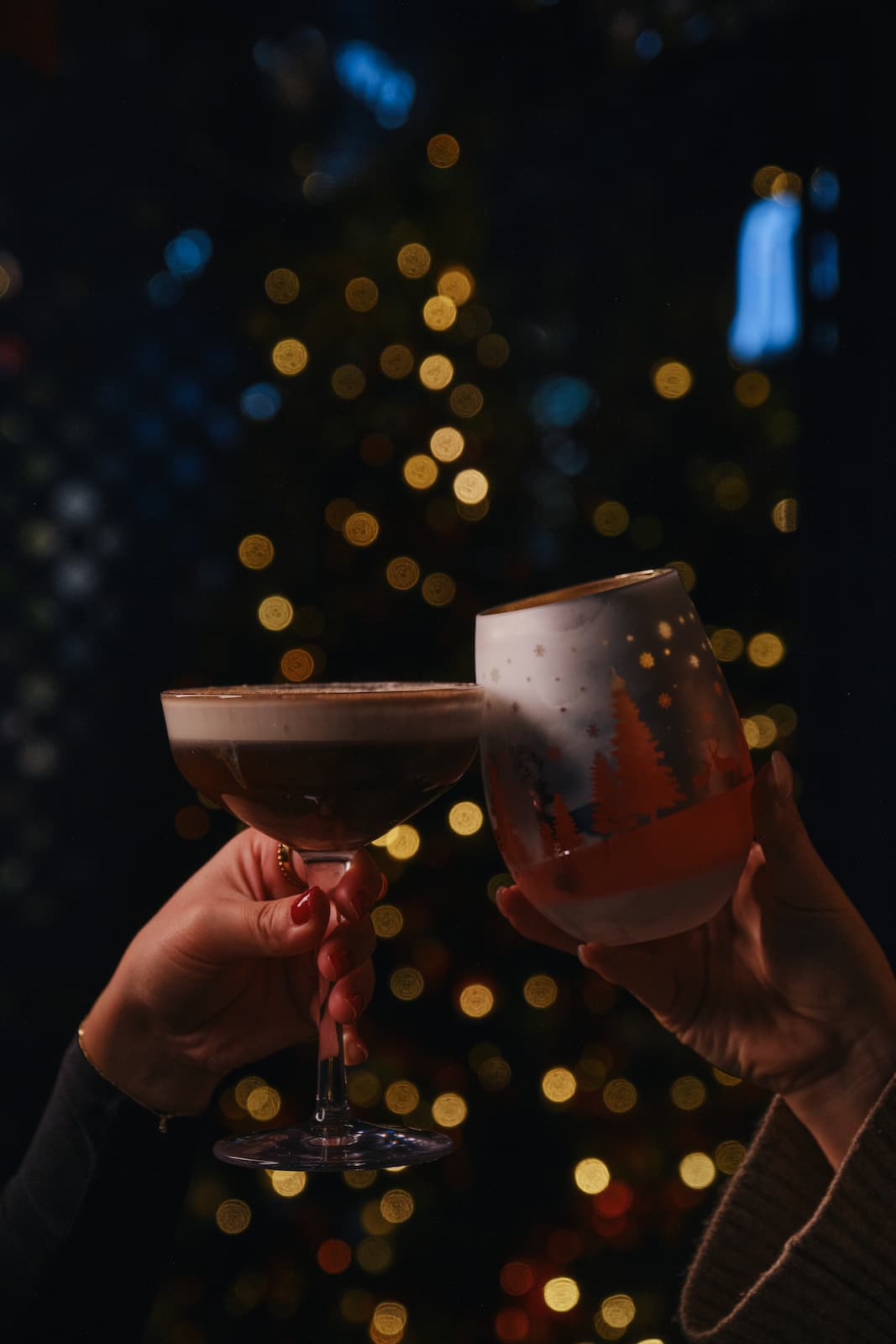 Two hands raising festive drinks, including a dark cocktail in a coupe glass and a decorated stemless glass, against a background of softly glowing Christmas lights.